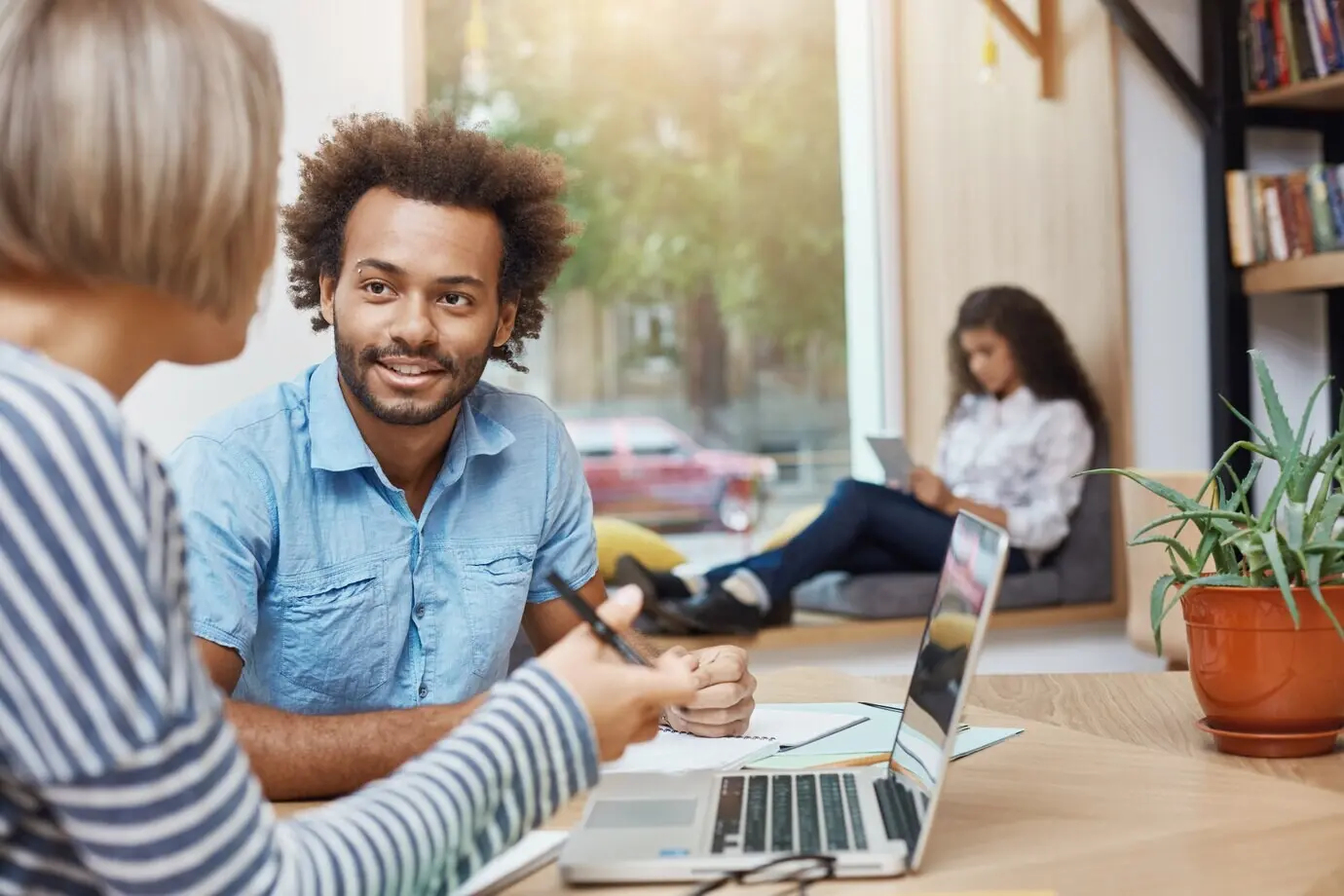 Крупный план молодого привлекательного темнокожего фрилансера с кудрявыми волосами в повседневной одежде, сидящего в библиотеке, разговаривающего с клиентом и показывающего на ноутбуке примеры своих работ.
